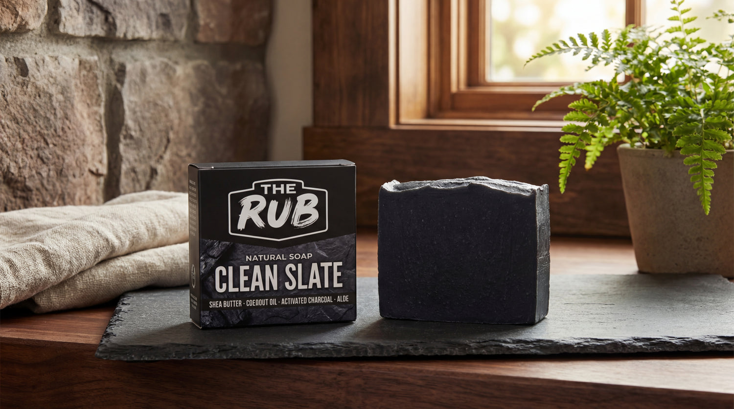 Clean Slate natural soap bar and box by The Rub displayed on a slate slab with a rolled towel and a stone wall backdrop.