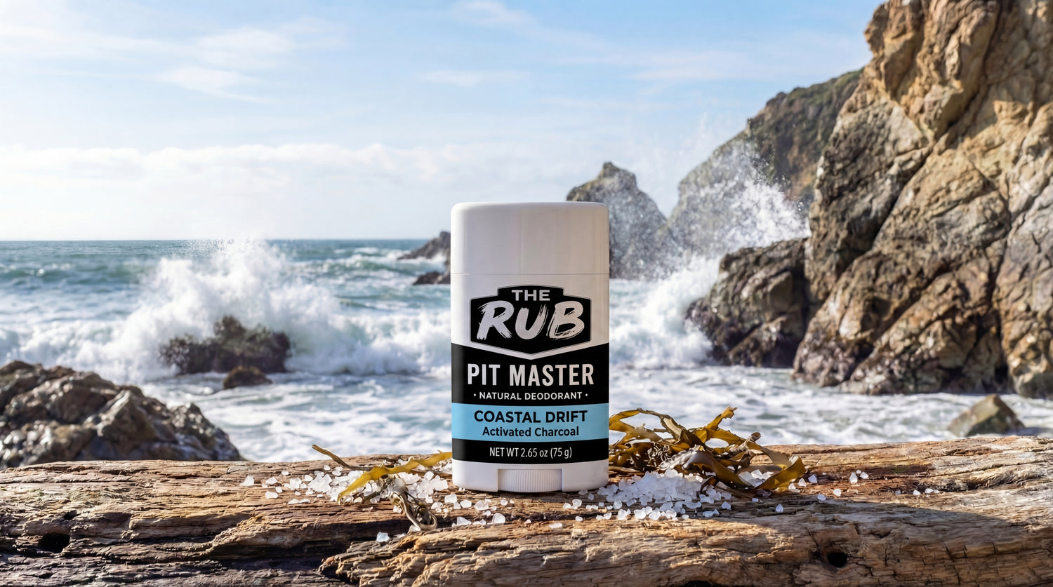 The Rub Coastal Drift natural deodorant sitting on driftwood with sea salt and seaweed against a rocky ocean coast.