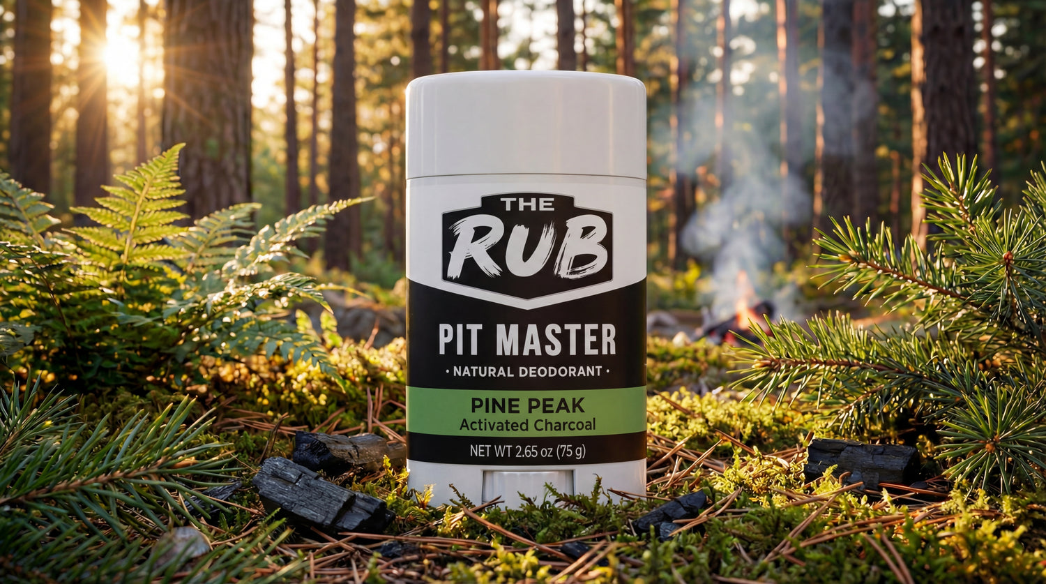The Rub Pine Peak natural deodorant stick sitting on a sunlit pine forest floor with charcoal and a distant campfire.