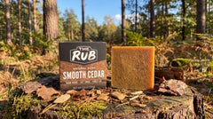 Smooth Cedar natural soap bar and box by The Rub displayed on a mossy tree stump in a sunlit pine forest.